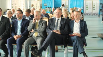 Einige Menschen sitzen in mehreren Stuhlreihen in einer Halle. 