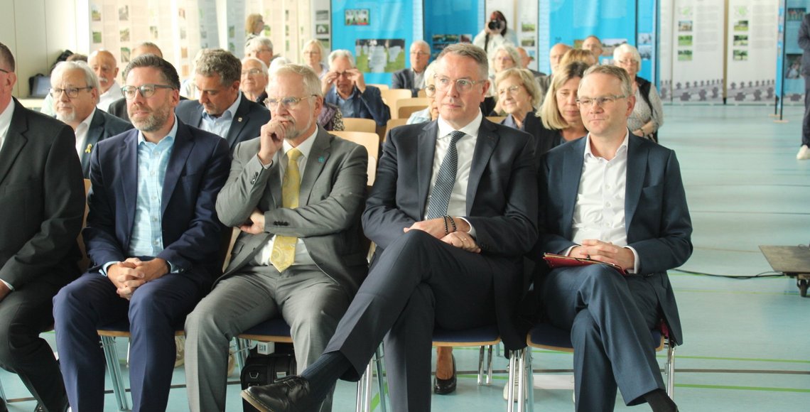 Einige Menschen sitzen in mehreren Stuhlreihen in einer Halle. 