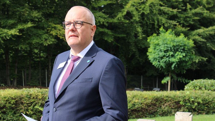 Ein Mann im Anzug spricht auf einem Friedhof. 
