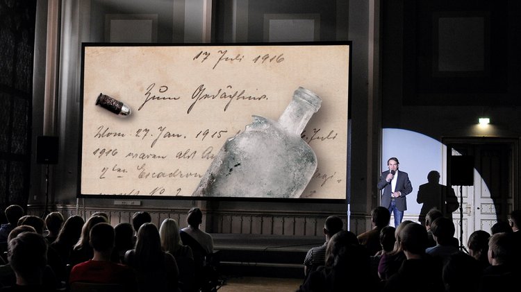 Eine große Leinwand vor Publikum, darauf ist ein Feldpostbrief und eine alte Flasche zu sehen. Neben der Leinwand steht ein Mann, der in ein Mikrofon spricht.