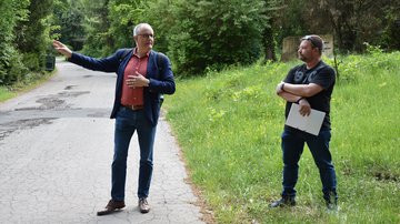 Zwei Männer stehen nebeneinander auf einem asphaltierten Weg bzw. auf dem Rasen daneben, einer von ihnen spricht und zeigt erklärend mit dem Arm nach rechts.