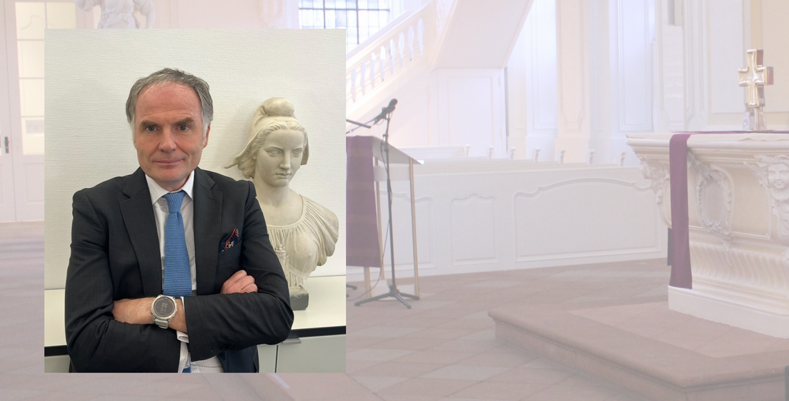 Ein Mann im Anzug, der in die Kamera schaut. Im Hintergrund der Innenraum einer Kirche.