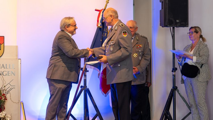 Ein Mann im Anzug verleiht ein Ehrenkreuz an einen Mann in Uniform und schüttelt dessen Hand