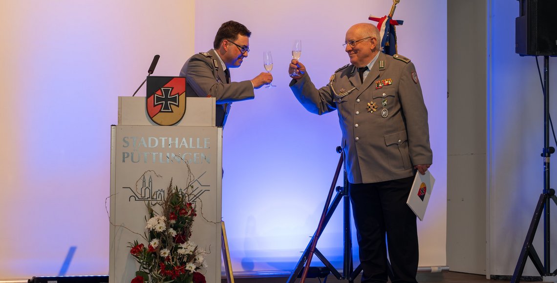 Zwei Männer in Uniform prosten sich auf einer kleinen Bühne mit Sektgläsern zu.