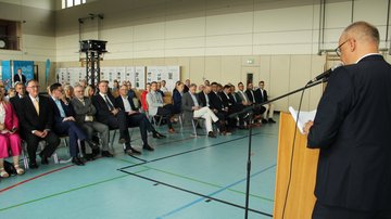 Ein Mann spricht am Rednerpult; vor ihm sitzen rund 50 Personen in Stuhlreihen in einer Halle. 