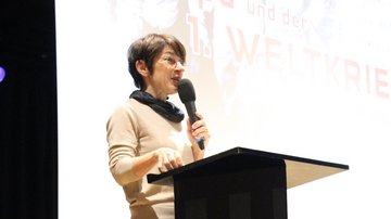 Eine Frau steht auf einer Bühne und spricht in ein Mikrofon.