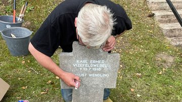 Ein Mann beugt sich von hinten über ein Grabkreuz und zieht die Schrift nach