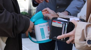 Eine Hand hält ein Portemonnaie, eine andere Hand hält eine Sammeldose vom Volksbund fest.