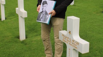 Ein Mann zeigt ein Schwarz-weiß-Foto eines Soldaten neben einem weißen Grabkreuz.
