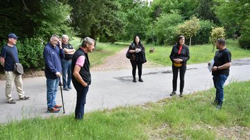 Sieben Personen stehen verteilt auf einem asphaltierten Weg und hören einem Redner zu