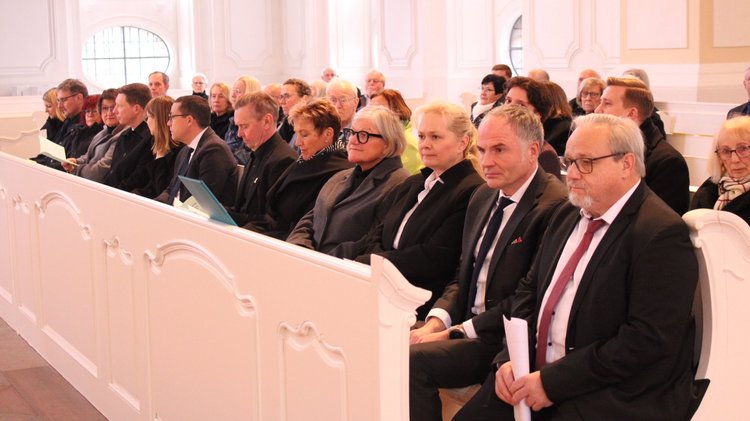 Rund fünfzehn gut gekleidete Personen sitzen nebeneinander in einer Kirchenbank.