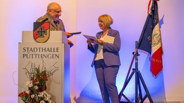 Ein Mann in Uniform und eine Frau stehen sich auf einer Bühne gegenüber und lachen.