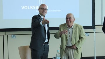 Ein jüngerer Mann und ein älterer Mann stehen lachend nebeneinander.