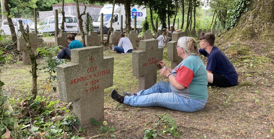 Mehrere Personen sitzen vor Kriegsgräbern und ziehen die Schrift mit einem Stift nach